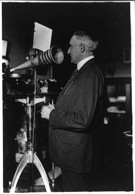 President Warren G. Harding, three-quarter length, speaking into a large funnel-shaped recording horn while making a voice recording, early 1920s.