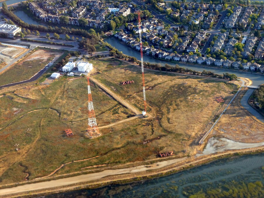 Aerial view of the KNBR 680 transmitter site on the bay marsh at the Belmont/Redwood City line, showing the top-loaded main tower and auxiliary tower